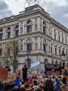 A crowd of protestors facing 10 Downing Street. A trans flag is waving in the wind in the middle of the crowd, and next to it a sign that states "respect our existence or expect our resistance".